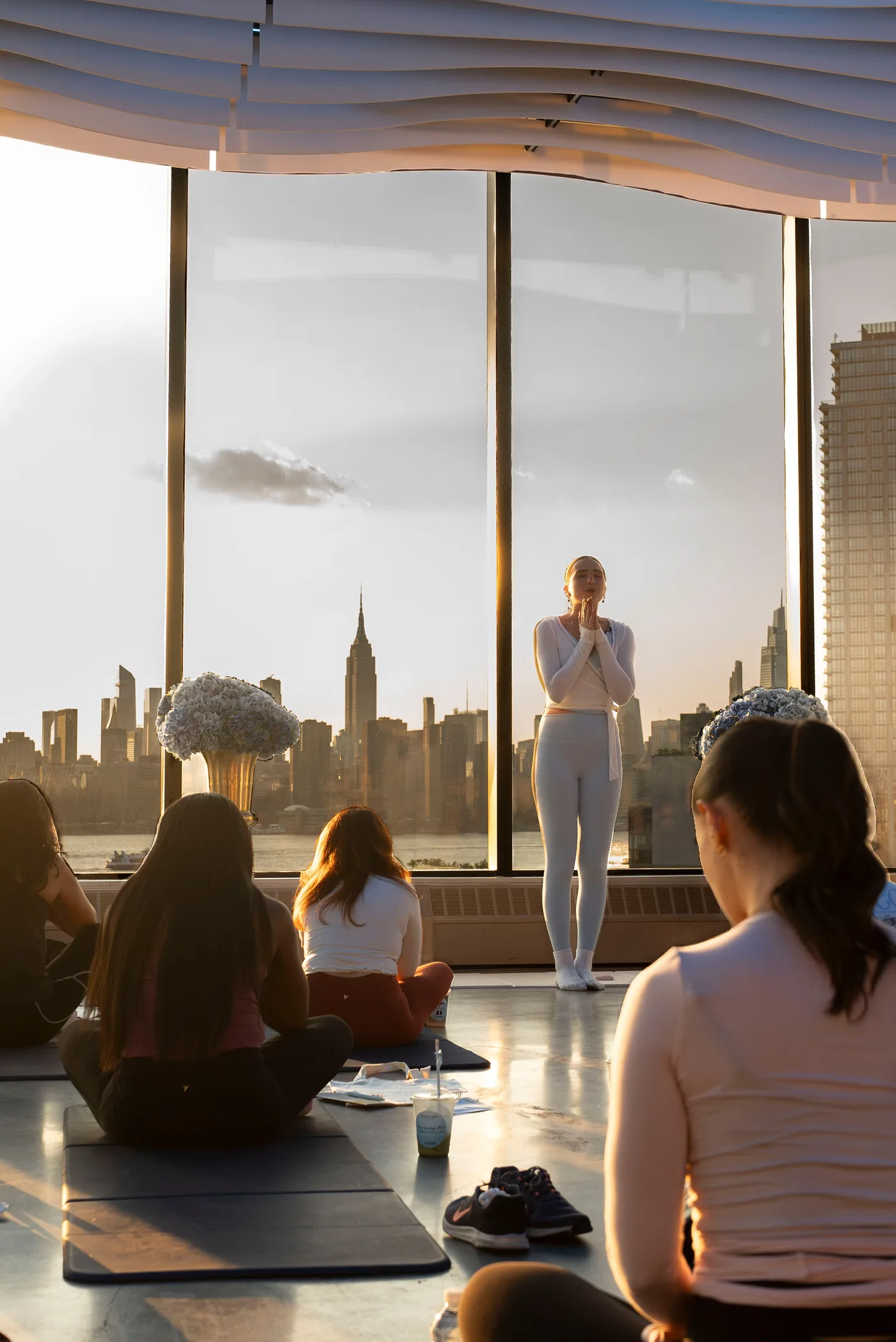 A wellness class instructor in white leads a sunset session at VESPER with the Manhattan skyline behind her in Brooklyn