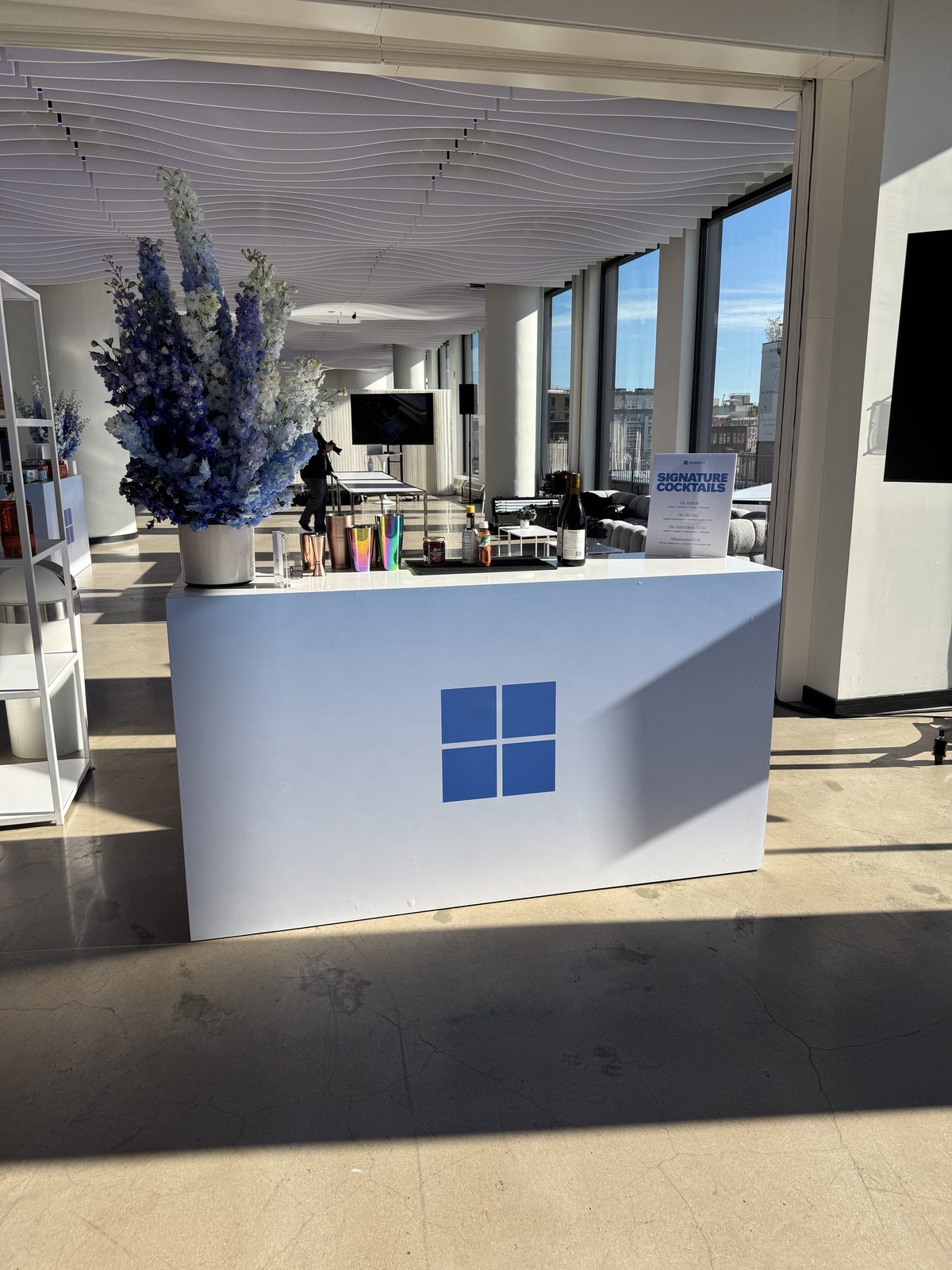 A Windows-branded bar with blue florals and signature cocktail signs in front of skyline windows at VESPER