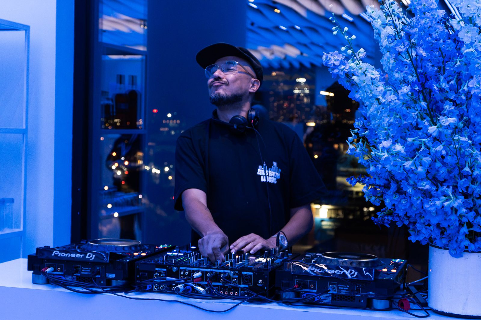 A DJ playing beside tall blue florals in front of the night skyline at VESPER