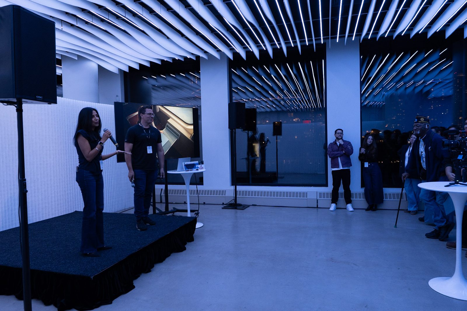 Presenters on a small stage with a screen and a standing crowd at VESPER